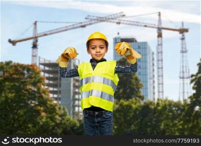 building and profession concept - smiling little boy in protective gloves, yellow safety vest and helmet showing his power over construction site background. little boy showing power over construction site