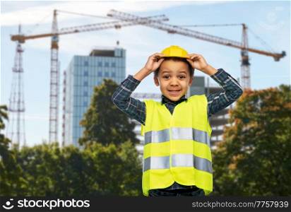 building and profession concept - smiling little boy in yellow safety vest and helmet over construction site background. little boy in safety vest over construction site