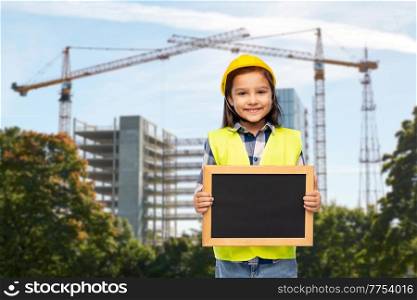 building and profession concept - smiling little girl in protective helmet and safety vest holding chalkboard over construction site background. little girl in construction helmet with chalkboard