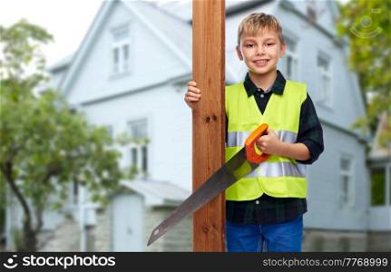 building, carpentry and profession concept - happy smiling little boy in protective helmet and safety vest with saw and wooden board over living house background. little boy in safety vest with saw and board