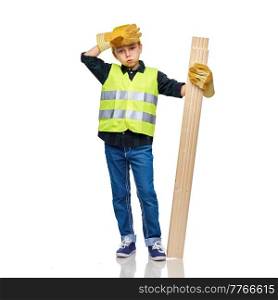 building, carpentry and profession concept - tired little boy in protective helmet, gloves and safety vest with wooden boards over white background. tired boy in protective helmet with wooden boards