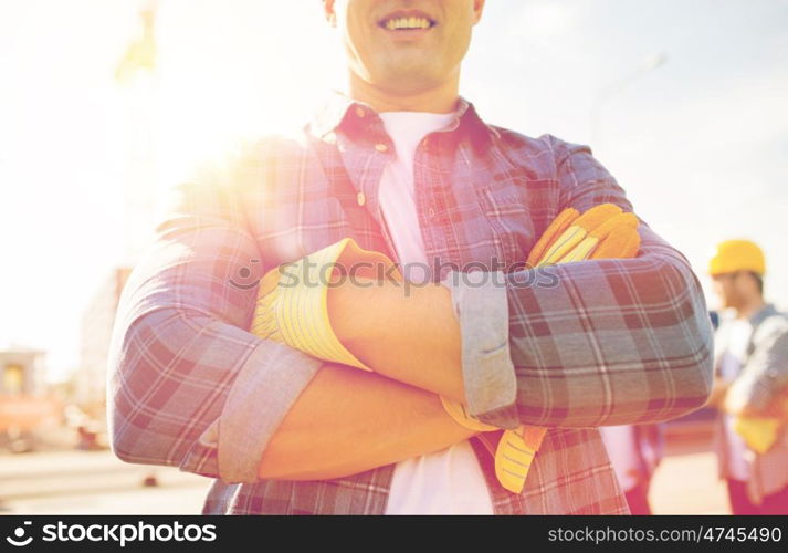 building, construction and people concept - close up of of smiling builder with crossed hands in gloves outdoors