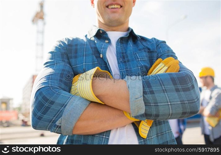 building, construction and people concept - close up of of smiling builder with crossed hands in gloves outdoors