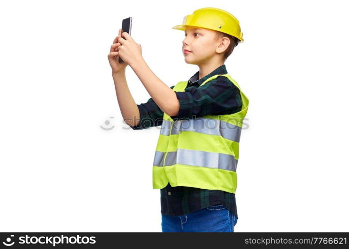 building, construction and profession concept - happy smiling little boy in protective helmet and safety vest taking selfie with smartphone over white background. little boy in helmet taking selfie with phone