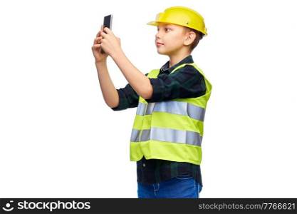 building, construction and profession concept - happy smiling little boy in protective helmet and safety vest taking selfie with smartphone over white background. little boy in helmet taking selfie with phone