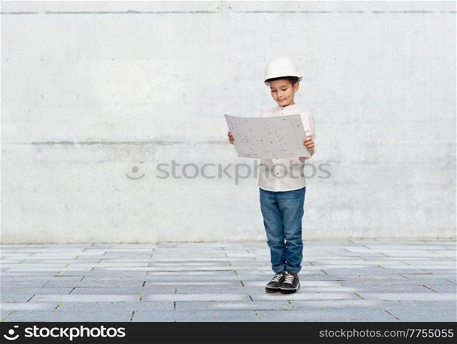 building, construction and profession concept - smiling little boy in helmet with blueprint over grey concrete wall background. little boy in construction helmet with blueprint