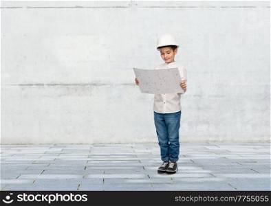 building, construction and profession concept - smiling little boy in helmet with blueprint over grey concrete wall background. little boy in construction helmet with blueprint