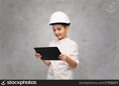 building, construction and profession concept - smiling little boy in helmet with tablet pc computer over grey concrete wall background. little boy in construction helmet with tablet pc
