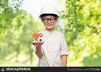 building, construction and profession concept - smiling little boy in helmet with toy house model over green natural background. little boy in construction helmet with house model