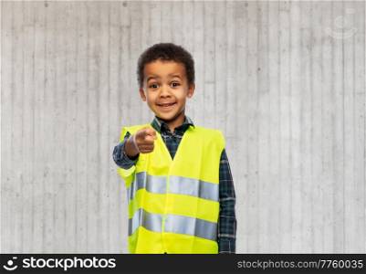 building, construction and profession concept - smiling little boy in yellow safety vest pointing finger to camera over grey concrete wall background. little boy in safety vest over white background