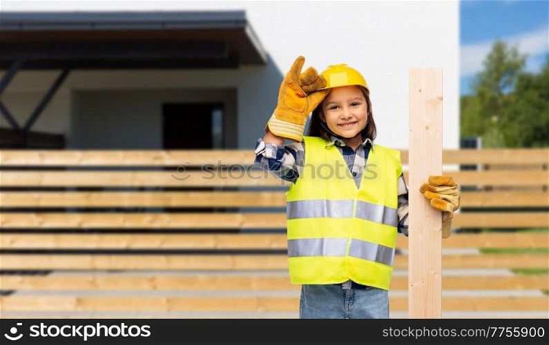 building, construction and profession concept - smiling little girl in protective helmet, gloves and safety and wooden board vest over house background. little girl in construction helmet with board