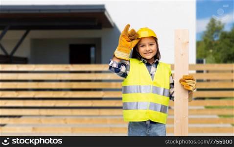 building, construction and profession concept - smiling little girl in protective helmet, gloves and safety and wooden board vest over house background. little girl in construction helmet with board