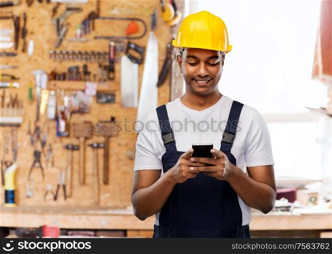 building, construction and technology concept - happy smiling indian worker or builder in helmet using smartphone over work tools on background. happy indian or builder in helmet using smartphone