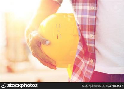 building, construction, protective gear and people concept - close up of builder hand holding yellow hardhat or helmet at construction site