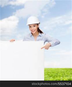 building, developing, consrtuction and architecture concept - young smiling businesswoman in helmet pointing finger to white blank board
