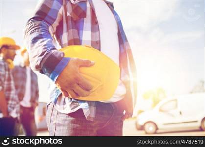 building, protective gear and people concept - close up of builder hand holding yellow hardhat or helmet at construction site