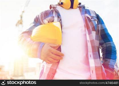 building, protective gear and people concept - close up of builder holding yellow hardhat or helmet outdoors at construction site