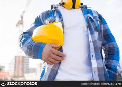 building, protective gear and people concept - close up of builder holding yellow hardhat or helmet outdoors at construction site