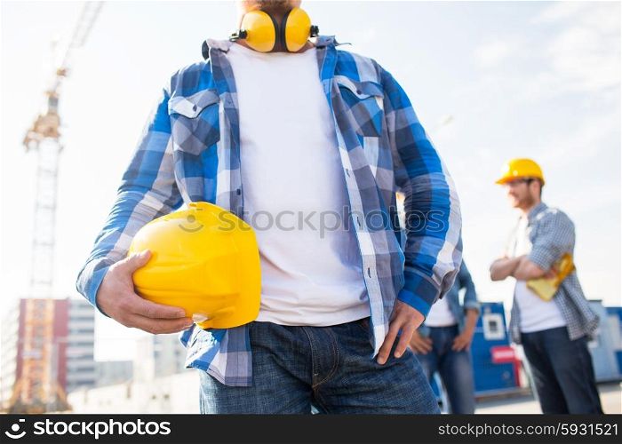building, protective gear and people concept - close up of builder holding yellow hardhat or helmet at construction site