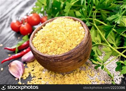 Bulgur groats - steamed wheat grains - in a clay bowl and a spoon on burlap napkin, tomatoes, hot peppers, garlic and parsley on black wooden board background