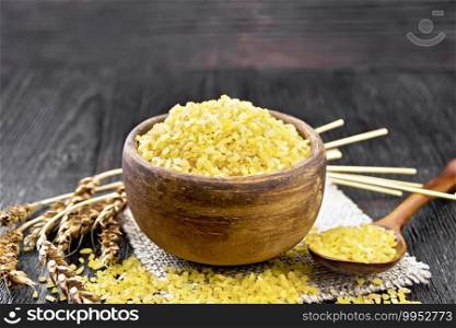 Bulgur groats - steamed wheat grains - in a clay bowl and a spoon on a burlap napkin, wheat ears on wooden board background