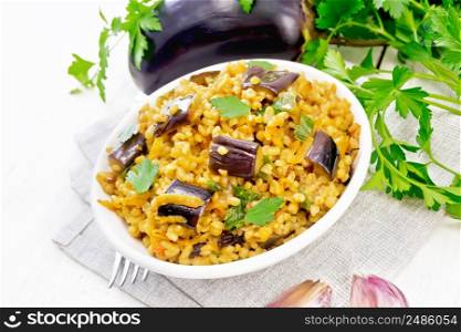 Bulgur stewed with eggplants, carrots, garlic and onions in a bowl on a towel against the background of light wooden board