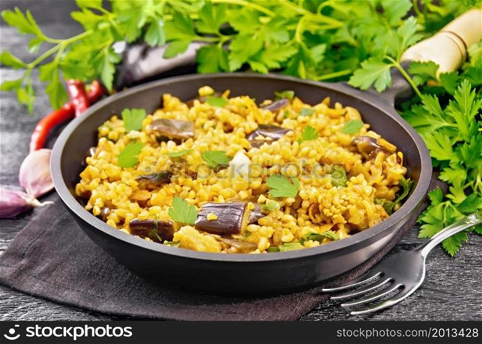 Bulgur stewed with eggplants, carrots, garlic and onions in a frying pan on a towel on wooden board background