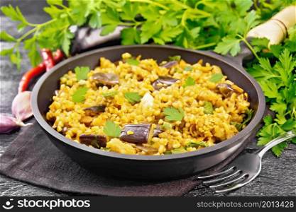 Bulgur stewed with eggplants, carrots, garlic and onions in a frying pan on a towel on wooden board background