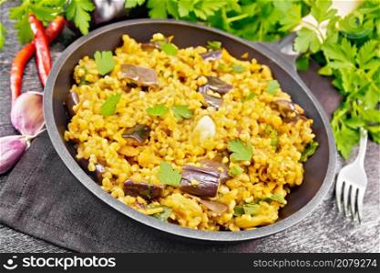 Bulgur stewed with eggplants, carrots, garlic and onions in a frying pan on a towel on black wooden board background