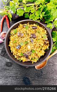 Bulgur stewed with eggplants, carrots, garlic and onions in a frying pan on a towel against the background of dark wooden board from above