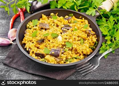 Bulgur stewed with eggplants, carrots, garlic and onions in a frying pan on a towel against the background of dark wooden board