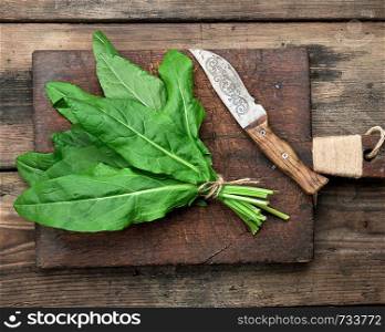 bunch of fresh green sorrel leaves and old brown cutting board on a wooden table, top view