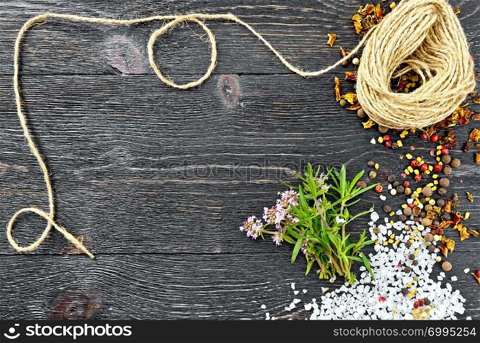 Bunch of fresh thyme with green leaves and pink flowers, salt, pepper, fenugreek seeds and a coil of twine against a black wooden board