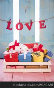 Bunch of multicolored presents in a white wooden crate on a pink table, with the word love tied to a string on a blue wooden fence, in the background.