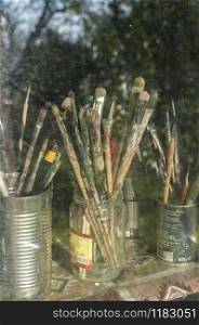 Bunch of old used grunge paint brushes in cans and jars behind window glass in artist studio