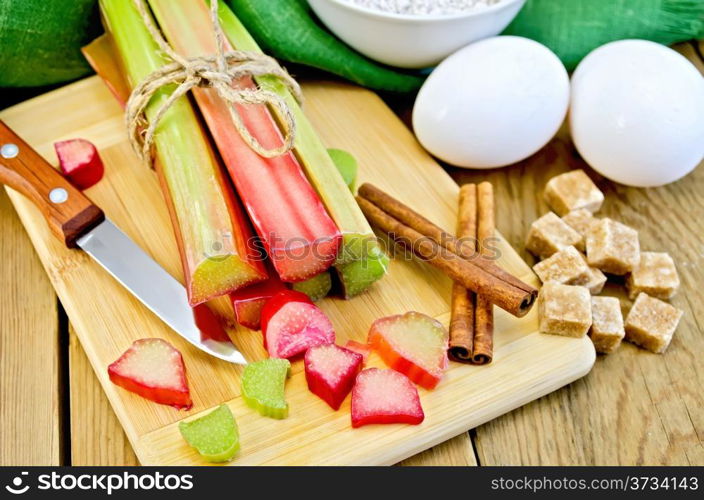 Bundle of stalks rhubarb, cut pieces of rhubarb, knife, sugar cubes, cinnamon, two eggs, flour, napkin on wooden board