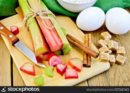 Bundle of stalks rhubarb, cut pieces of rhubarb, knife, sugar cubes, cinnamon, two eggs, flour, napkin on wooden board