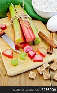 Bundle of stalks rhubarb, cut pieces of rhubarb, knife, sugar cubes, cinnamon, eggs, flour, cloth on a wooden board