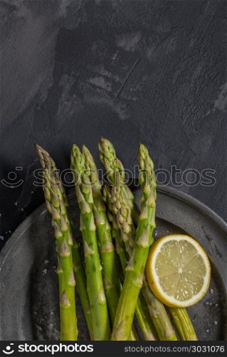 Bundle of young raw uncooked organic green asparagus with sliced. Bundle of young raw uncooked organic green asparagus with sliced lemon and sea salt over black texture background. Top view. Healthy eating