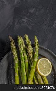 Bundle of young raw uncooked organic green asparagus with sliced. Bundle of young raw uncooked organic green asparagus with sliced lemon and sea salt over black texture background. Top view. Healthy eating