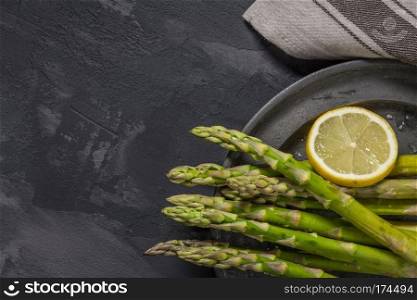 Bundle of young raw uncooked organic green asparagus with sliced lemon and sea salt over black texture background. Top view. Healthy eating