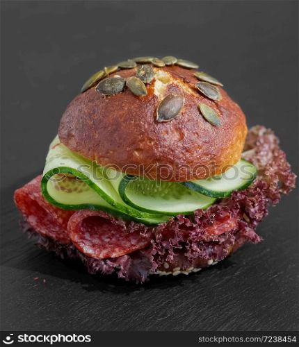 Burger with salami, lettuce and cucumber. Bun with sesame seeds and pumpkin seeds. Quick breakfast. Black stone background. Close-up.