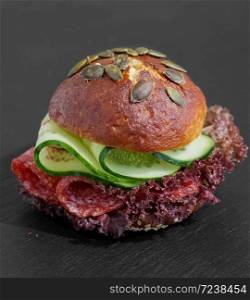 Burger with salami, lettuce and cucumber. Bun with sesame seeds and pumpkin seeds. Quick breakfast. Black stone background. Close-up.