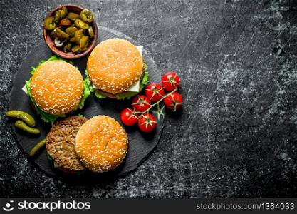 Burgers with tomatoes and gherkins on a stone Board. On black rustic background. Burgers with tomatoes and gherkins on a stone Board.