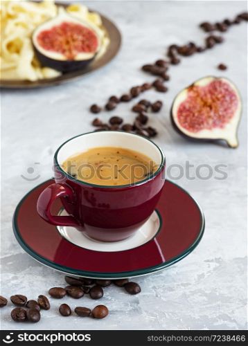 Burgundy cup with freshly brewed espresso on a gray background. Roasted coffee beans are located around a cup of coffee. In the background is fresh figs and homemade cheese.. Burgundy cup with freshly brewed espresso on a gray background. Roasted coffee beans are located around a cup of coffee. In the background is fresh figs and homemade cheese. Close-up.