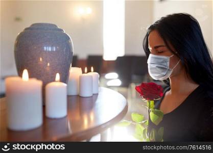 burial, people and mourning concept - sad woman wearing face protective medical mask for protection from virus disease with red rose and cinerary urn at funeral in church. woman in mask with rose at funeral in church