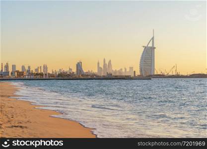 Burj Al Arab in Jumeirah Island or boat building with waves on sea beach, Dubai Downtown skyline, United Arab Emirates or UAE. Financial district in urban city. Skyscrapers at sunset.