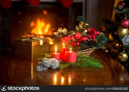 Burning candles, Christmas wreath and golden gift box on table next to burning fireplace at living room. Image with shallow depth of focus.