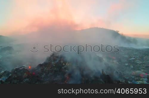 Burning pile of garbage at dump site toxic smoke rises into the air against setting sun