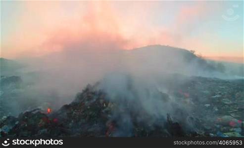 Burning pile of garbage at dump site toxic smoke rises into the air against setting sun